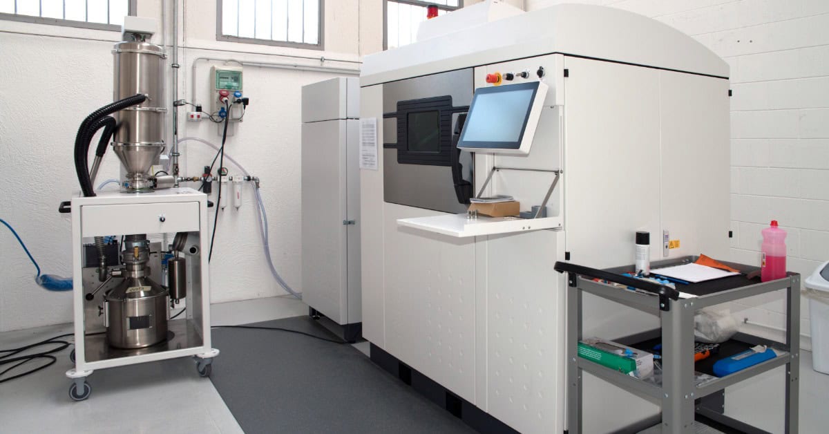 A large white industrial machine features a display touchscreen and a small window to view the progress.