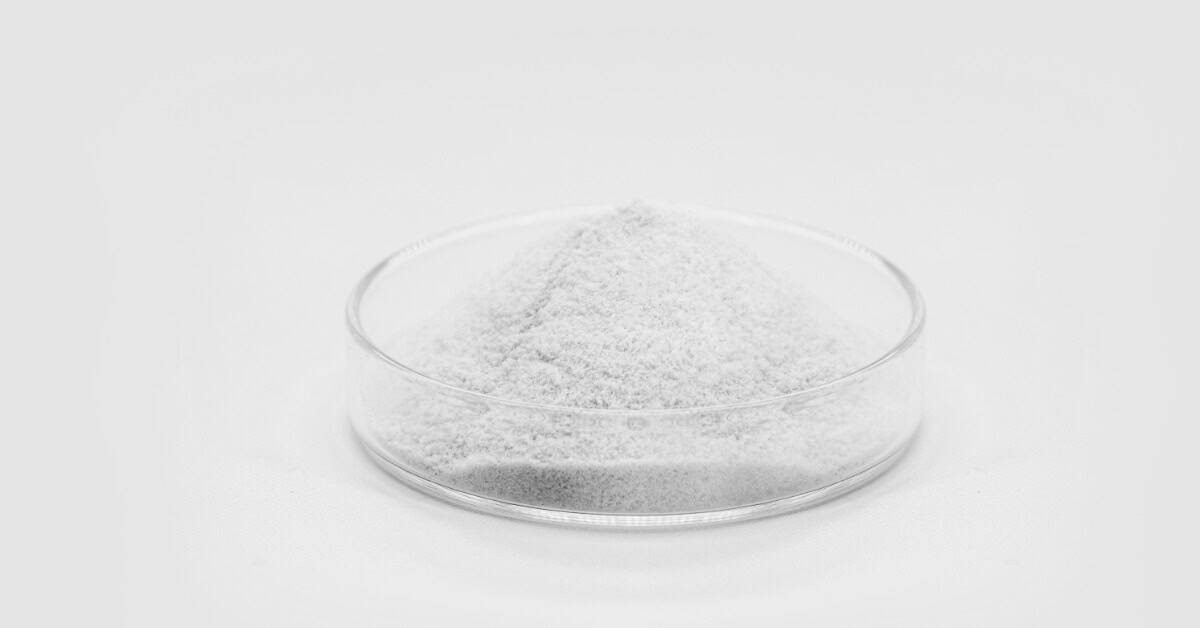 A small, shallow glass dish contains a white powder. The dish sits alone on a stark, white background.