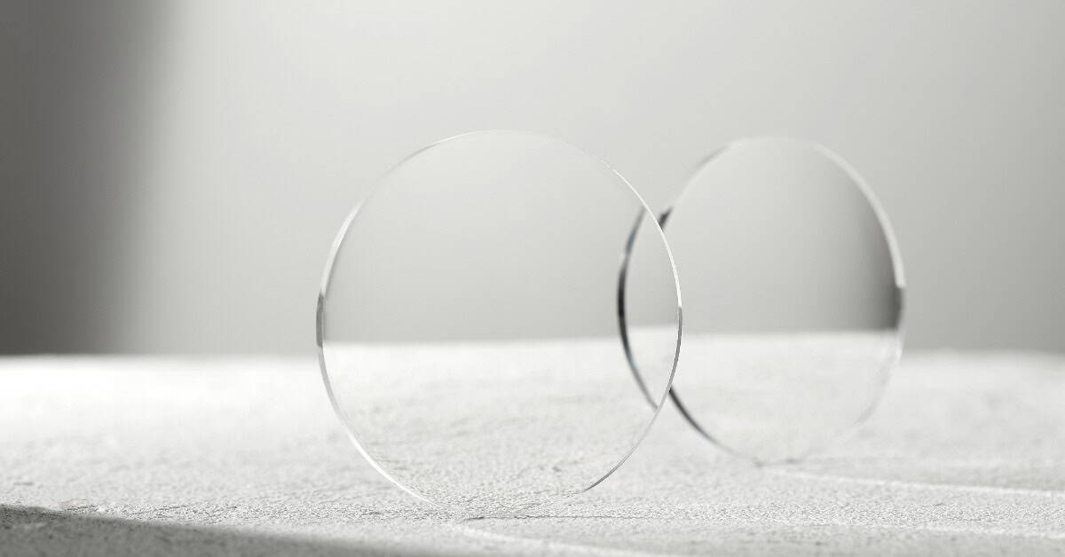 Two lenses for glasses are sitting on their edge on a gray-white surface. The background is gray and blurry.