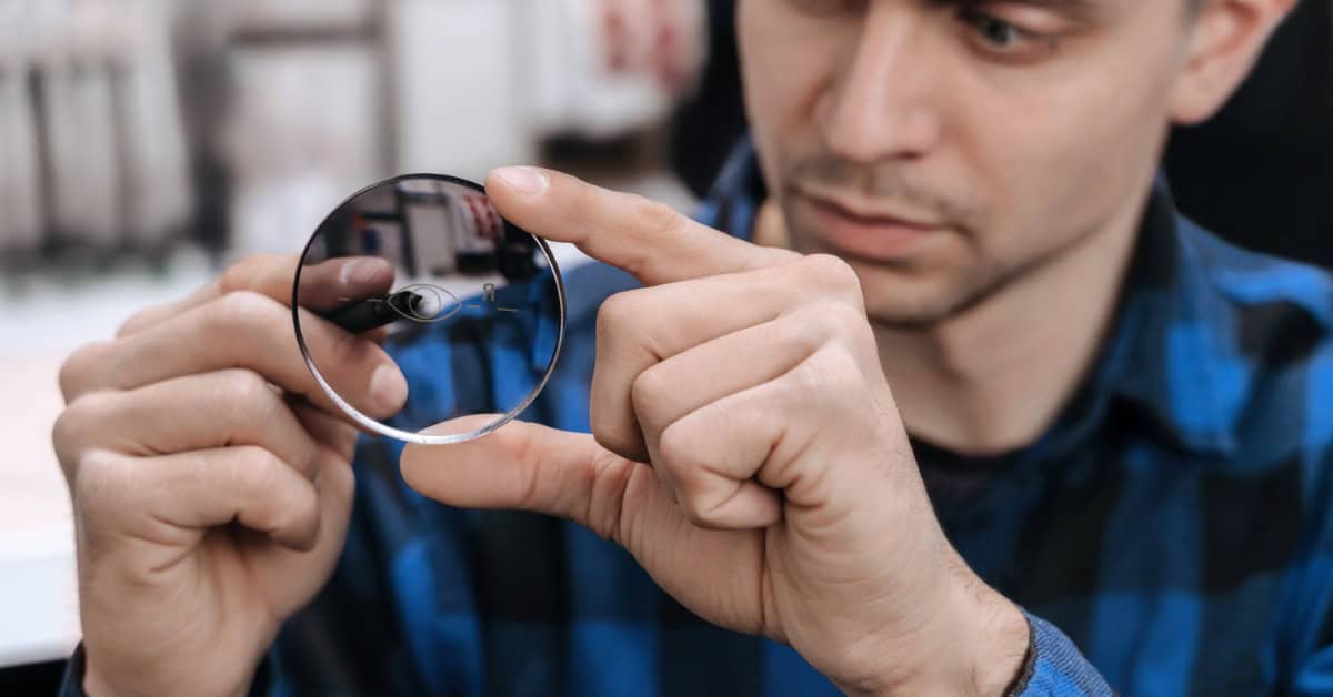 A close-up view shows a man wearing a black and blue plaid shirt working on a lens with a special tool.
