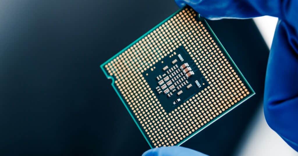 A close-up view shows a person wearing a blue latex glove holding a semiconductor microchip against a black surface.
