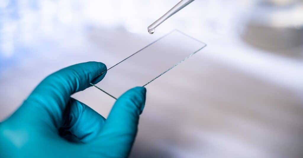 A close-up view shows a person wearing teal latex gloves using a pipette to apply a liquid to a fused quartz slide.