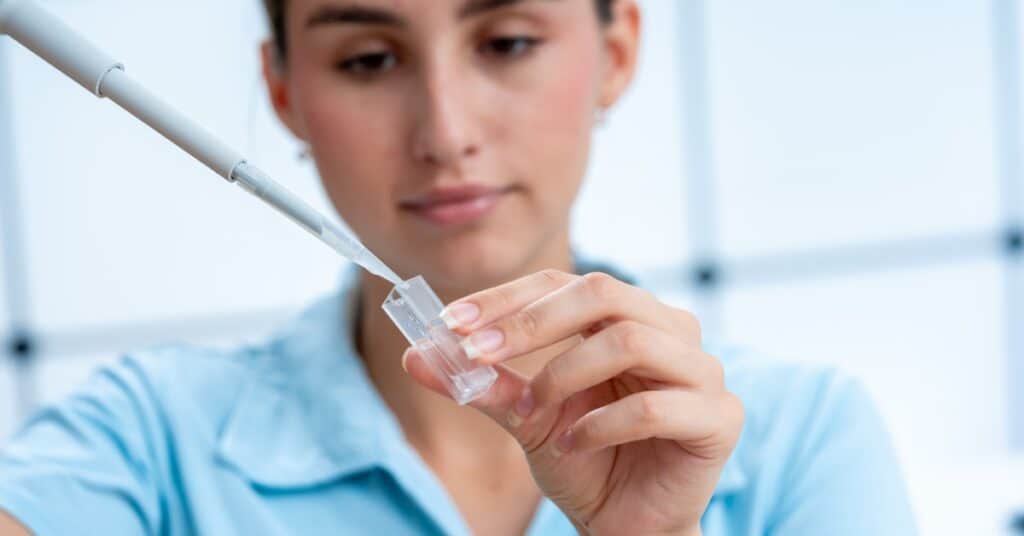 A woman wearing a baby blue shirt is using a large pipette to place liquid into a fused quartz cuvette.